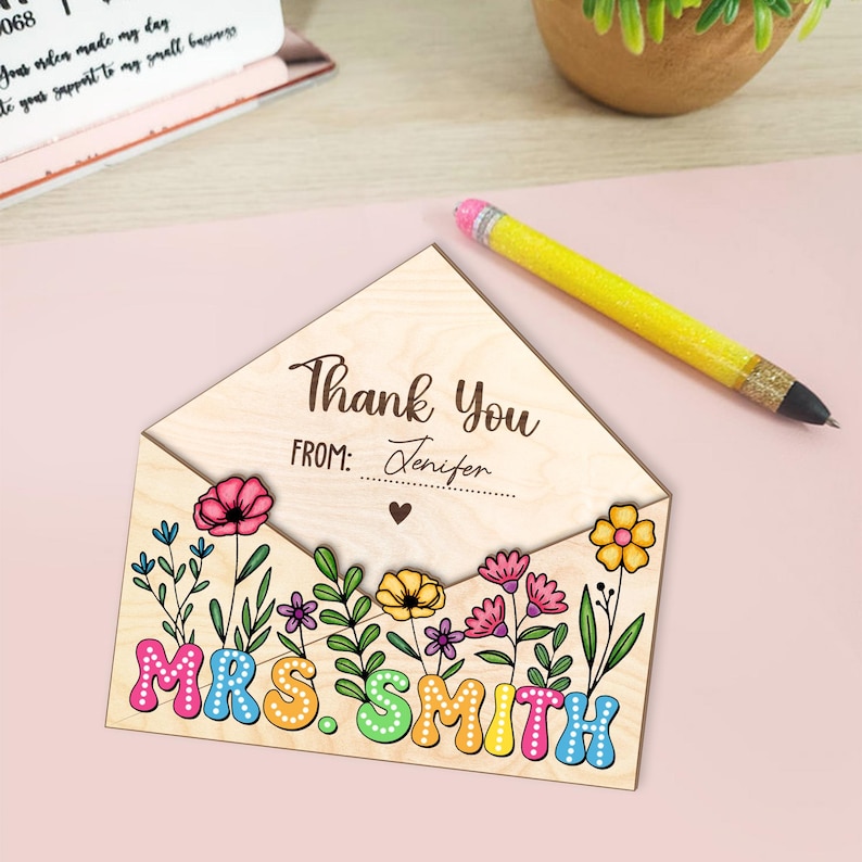 May include: A wooden thank you card with a floral design. The card is shaped like an envelope and has the text "Thank You" and "From: Jenifer" on the front. The back of the card has the text "MRS. SMITH" in colorful letters.