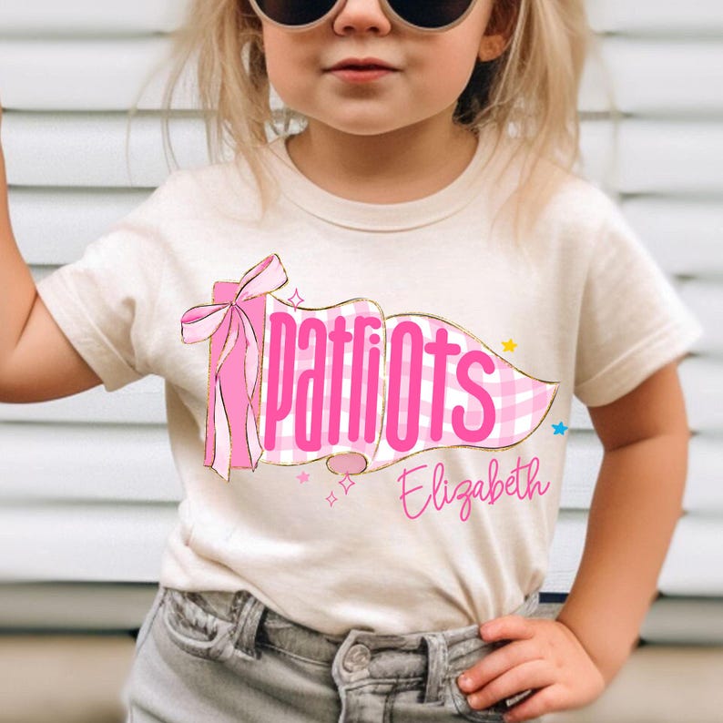 May include: A cream-colored short-sleeve t-shirt featuring a graphic of a pink gingham pennant flag with a pink bow. The text "Patriots" is printed in bold pink letters across the flag, with the name "Elizabeth" written below in pink script.