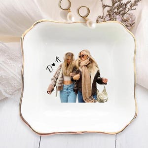 Pu&ograve; includere: Un vassoio portagioie in ceramica bianca con bordo dorato, con una foto di due donne. Il vassoio ha una forma quadrata con bordi smerlati. Due orecchini di perle e fiori secchi sono posizionati vicino al vassoio.