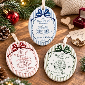 Op de afbeelding: Drie ovale keramische ornamenten met gestreepte randen en linten. Elk bevat de tekst The Year We Got Engaged, Laura & Rylan en 2026. Eén heeft blauwe strepen en champagneglazen, één rode strepen en hartvormige medaillons, en één groene strepen en een auto.