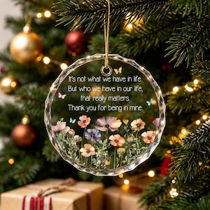 Op de afbeelding: Een rond, gefacetteerd ornament van helder glas met een gouden ophangkoord. Het oppervlak toont een kleurrijke illustratie van een bloemenweide met kleine vlinders en de tekst: It's not what we have in life. But who we have in our life, that really matters. Thank you for being in mine.