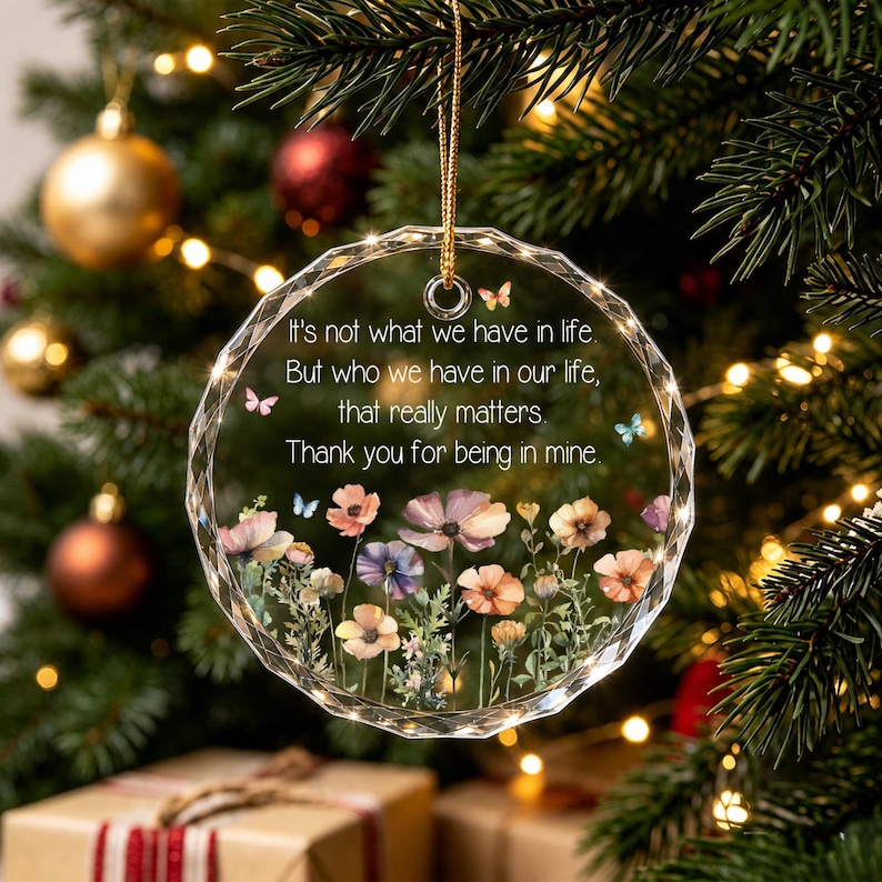 Op de afbeelding: Een rond, gefacetteerd ornament van helder glas met een gouden ophangkoord. Het oppervlak toont een kleurrijke illustratie van een bloemenweide met kleine vlinders en de tekst: It's not what we have in life. But who we have in our life, that really matters. Thank you for being in mine.