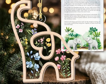 Op de afbeelding: Houten ornament in de vorm van een olifant met een bloemenontwerp. Het ornament heeft een helder midden gevuld met kleurrijke bloemen. Een kaart met tekst en een illustratie van twee olifanten is zichtbaar.