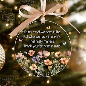 Op de afbeelding: Een cirkelvormig, transparant acryl ornament met een lichtroze strik aan de bovenkant. Het ornament toont een bedrukte bloemenweide met kleurrijke wilde bloemen en kleine vlinders, samen met de tekst: It's not what we have in life. But who we have in our life, that really matters. Thank you for being in mine.
