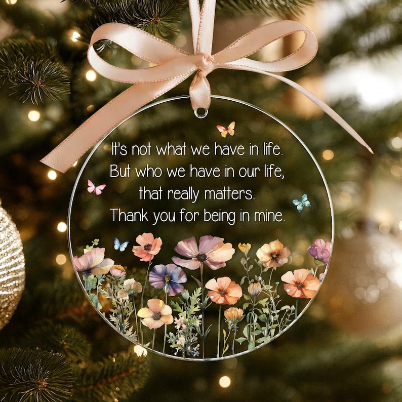 Op de afbeelding: Een cirkelvormig, transparant acryl ornament met een lichtroze strik aan de bovenkant. Het ornament toont een bedrukte bloemenweide met kleurrijke wilde bloemen en kleine vlinders, samen met de tekst: It's not what we have in life. But who we have in our life, that really matters. Thank you for being in mine.