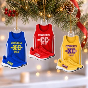 May include: Three flat Christmas ornaments shaped like tank tops and running shoes. The blue, red, and yellow ornaments feature the text COWGIRLS, XC or CC, KYLIE, and 2026. Each ornament is suspended by a gold string against a blurred tree background.