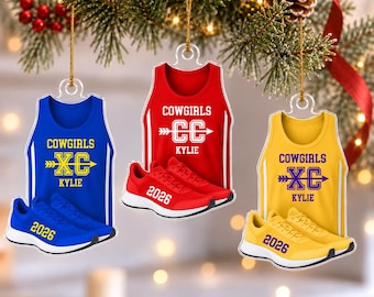 May include: Three flat Christmas ornaments shaped like tank tops and running shoes. The blue, red, and yellow ornaments feature the text COWGIRLS, XC or CC, KYLIE, and 2026. Each ornament is suspended by a gold string against a blurred tree background.