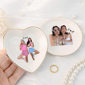 Pu&ograve; includere: Due portagioie in ceramica, uno a forma di cuore e uno rotondo, con bordi dorati. Il piatto a forma di cuore presenta una foto di due donne, le iniziali "H ❤️ A" e un anello d'oro. Il piatto rotondo ha una foto di due donne e le iniziali "H ❤️ A".