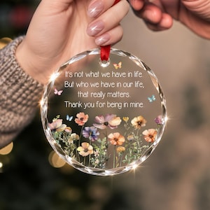 Op de afbeelding: Een rond, doorzichtig acryl ornament met gefacetteerde randen en een rood lint om op te hangen. In het midden staat de tekst: It's not what we have in life. But who we have in our life, that really matters. Thank you for being in mine. Onder de tekst staat een kleurrijke illustratie van diverse wilde bloemen en kleine vlinders.