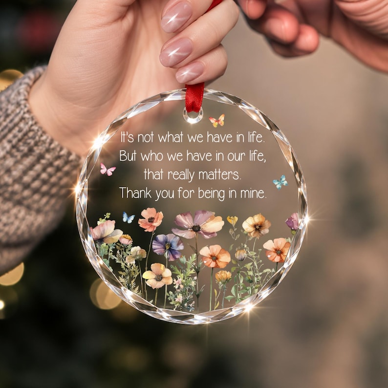 Op de afbeelding: Een rond, doorzichtig acryl ornament met gefacetteerde randen en een rood lint om op te hangen. In het midden staat de tekst: It's not what we have in life. But who we have in our life, that really matters. Thank you for being in mine. Onder de tekst staat een kleurrijke illustratie van diverse wilde bloemen en kleine vlinders.