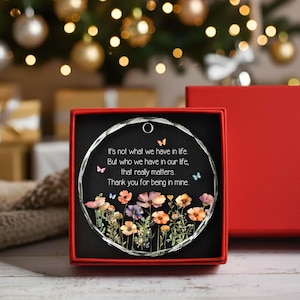 Op de afbeelding: Een rond glazen ornament in een rode geschenkdoos. Het ornament heeft een zwarte achtergrond met een kleurrijke illustratie van wilde bloemen onderaan, kleine vlinders en de tekst: It's not what we have in life, But who we have in our life, that really matters. Thank you for being in mine.