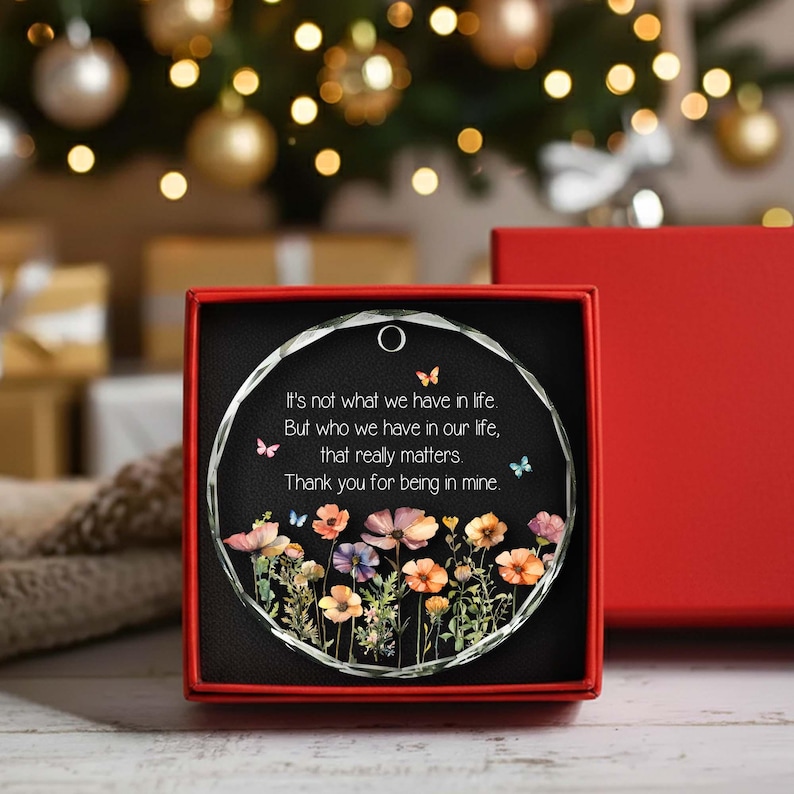 Op de afbeelding: Een rond glazen ornament in een rode geschenkdoos. Het ornament heeft een zwarte achtergrond met een kleurrijke illustratie van wilde bloemen onderaan, kleine vlinders en de tekst: It's not what we have in life, But who we have in our life, that really matters. Thank you for being in mine.