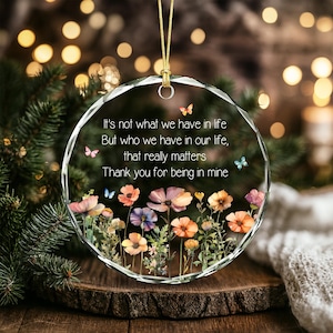 Op de afbeelding: Een rond ornament van helder glas met een gefacetteerde rand en een gouden ophangkoord. Het oppervlak toont een bedrukte bloementuin met kleurrijke wilde bloemen en kleine vlinders, samen met de tekst: It's not what we have in life But who we have in our life, that really matters Thank you for being in mine.