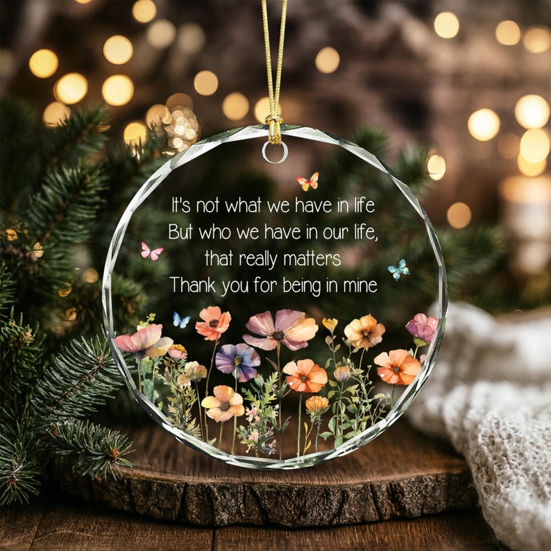 Op de afbeelding: Een rond ornament van helder glas met een gefacetteerde rand en een gouden ophangkoord. Het oppervlak toont een bedrukte bloementuin met kleurrijke wilde bloemen en kleine vlinders, samen met de tekst: It's not what we have in life But who we have in our life, that really matters Thank you for being in mine.