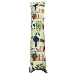 May include: A yellow cat-themed catnip toy in the shape of a long pillow. The pillow is covered in a fabric with a repeating pattern of cats, books, and plants on shelves. The text on the books includes "Cat & Kitten", "The Cat", and "Cat & Kitten".