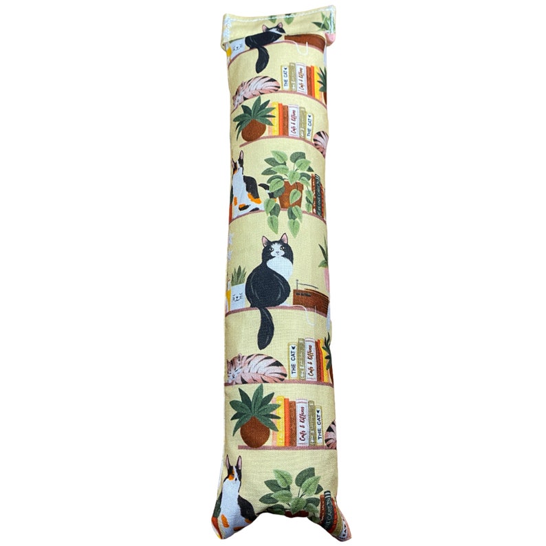May include: A yellow cat-themed catnip toy in the shape of a long pillow. The pillow is covered in a fabric with a repeating pattern of cats, books, and plants on shelves. The text on the books includes "Cat & Kitten", "The Cat", and "Cat & Kitten".