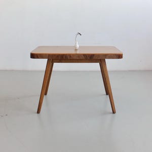 Op de afbeelding: Kleine, rechthoekige houten tafel met afgeronde hoeken en schuine poten. Het tafelblad is lichtbruin met een zichtbaar houtnerfpatroon. Een kleine witte vaas met een takje gedroogde bloemen staat erop.