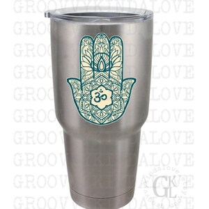 Puede incluir: Un vaso de acero inoxidable con un dise&ntilde;o de mano de hamsa de color azul verdoso y blanco con un s&iacute;mbolo om en el centro.