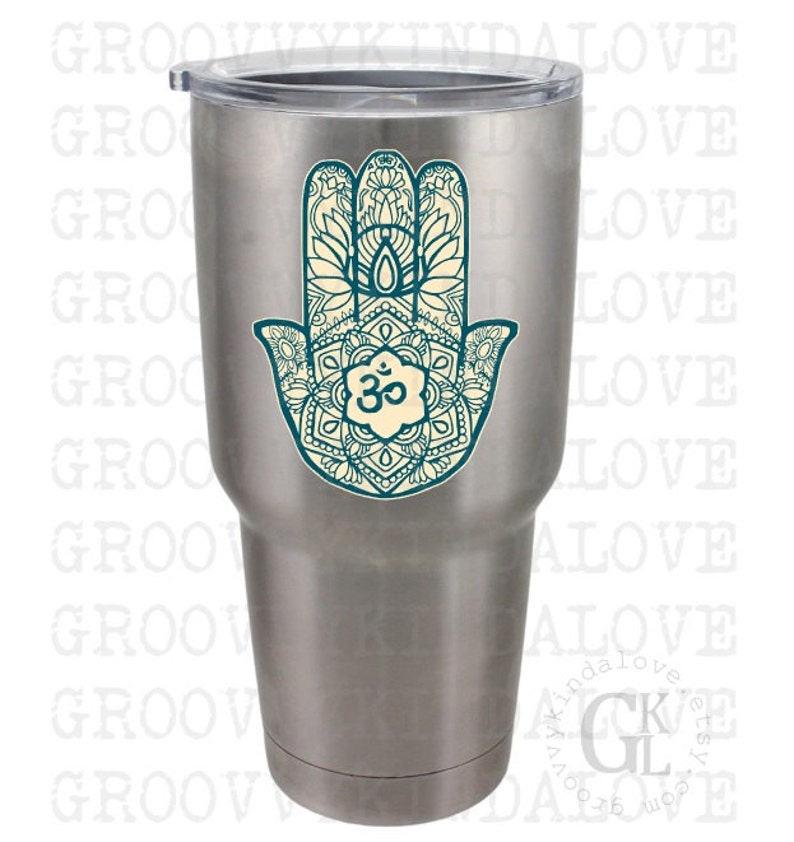 Puede incluir: Un vaso de acero inoxidable con un dise&ntilde;o de mano de hamsa de color azul verdoso y blanco con un s&iacute;mbolo om en el centro.