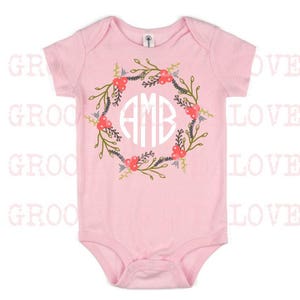 May include: Pink baby onesie with a white floral wreath and the monogram "AMB" in the center.