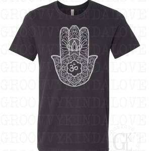 Puede incluir: Una camiseta gris oscuro con un dise&ntilde;o de mandala blanco de la mano de Hamsa, un s&iacute;mbolo de protecci&oacute;n y buena suerte. El mandala presenta detalles intrincados y el s&iacute;mbolo Om en el centro.