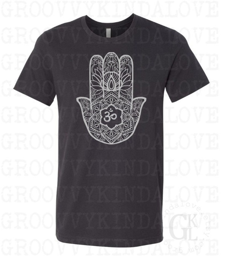 Puede incluir: Una camiseta gris oscuro con un dise&ntilde;o de mandala blanco de la mano de Hamsa, un s&iacute;mbolo de protecci&oacute;n y buena suerte. El mandala presenta detalles intrincados y el s&iacute;mbolo Om en el centro.
