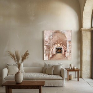 May include: A square canvas painting depicting a stone archway with a hanging chandelier and a dark interior passage. The artwork features textured brushstrokes in shades of beige, cream, and brown, displayed on a neutral wall above a sofa.