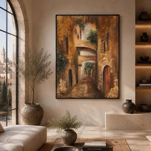 May include: A large rectangular framed oil painting depicting a narrow Mediterranean street scene with stone arches, textured golden-brown walls, and green foliage. The artwork features a dark wooden frame and is displayed on a neutral wall.