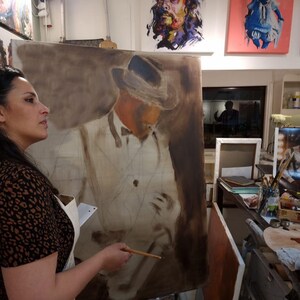 May include: A large rectangular canvas featuring an oil painting in progress of a figure wearing a hat and a suit. The painting uses a monochromatic brown and tan color palette to define the subject's form, with visible brushstrokes and outlines.