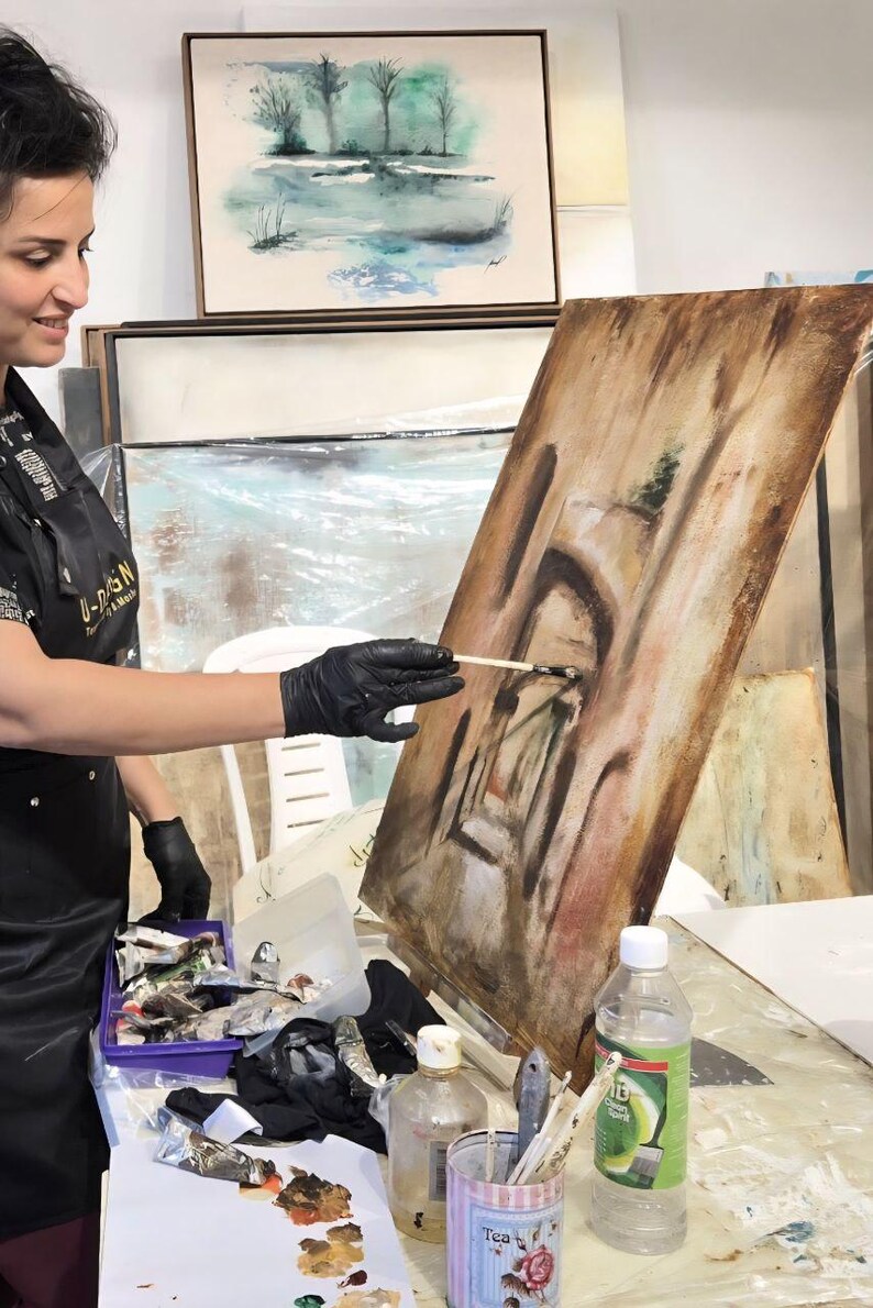 May include: A rectangular canvas on an easel featuring an abstract brown and tan painting with dark brushstrokes. The artist wears a black apron and black gloves while holding a paintbrush to the canvas. Paint tubes and a clear bottle are nearby.