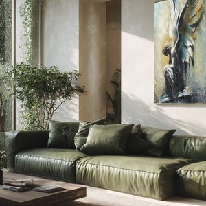Op de afbeelding: Een groot rechthoekig wandkunstwerk met een textuurrijk schilderij van een gevleugelde figuur in blauwe, gouden en witte tinten. Het kunstwerk hangt aan een lichte muur boven een lage olijfgroene leren bank met meerdere kussens.