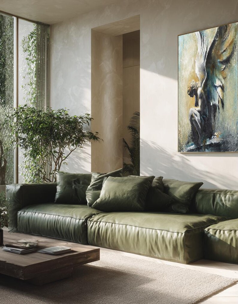 Op de afbeelding: Een groot rechthoekig wandkunstwerk met een textuurrijk schilderij van een gevleugelde figuur in blauwe, gouden en witte tinten. Het kunstwerk hangt aan een lichte muur boven een lage olijfgroene leren bank met meerdere kussens.