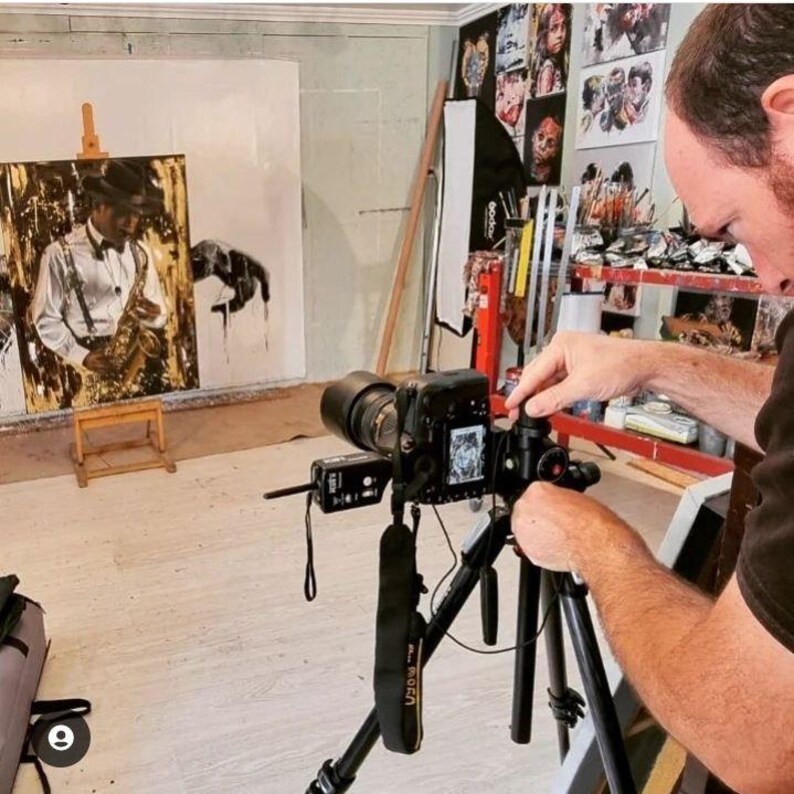 May include: A black digital camera mounted on a tripod, positioned to capture a large painting on an easel. The painting depicts a person in a hat and suspenders playing a saxophone. The scene is set in an art studio with various supplies.