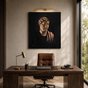 Op de afbeelding: Een vierkant wandschilderij met een portret van een man met krullend haar en een baard tegen een zwarte achtergrond, opgehangen boven een donkerhouten bureau met een bruine lederen bureaustoel, een messing lamp, een laptop en een kleine zwarte kom.