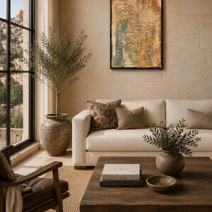 Pu&ograve; includere: Un soggiorno con un divano color crema con cuscini marroni e fantasia, un tavolino in legno scuro con un libro intitolato Jerusalem, un grande ulivo in vaso e un dipinto astratto strutturato su una parete in pietra beige.