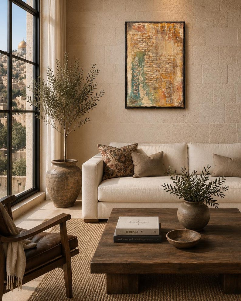 Pu&ograve; includere: Un soggiorno con un divano color crema con cuscini marroni e fantasia, un tavolino in legno scuro con un libro intitolato Jerusalem, un grande ulivo in vaso e un dipinto astratto strutturato su una parete in pietra beige.