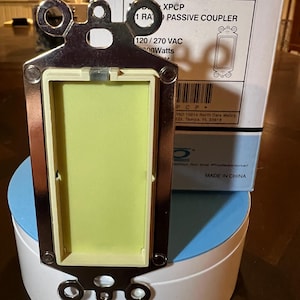 Puede incluir: Una placa de pared eléctrica plateada con un inserto de plástico blanco. La placa tiene tres orificios de montaje y una etiqueta que dice "XPCP 1 RA PASSIVE COUPLER 120/270 VAC Watts".