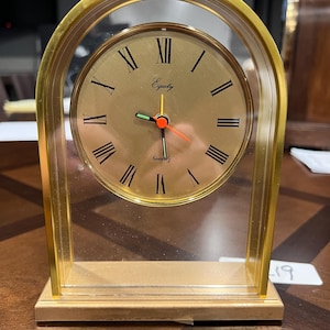 May include: A gold-coloured quartz clock with a clear acrylic frame. The clock face is gold with black Roman numerals and the word "Equity". The hands are black, green, and orange. The clock has a rectangular base.