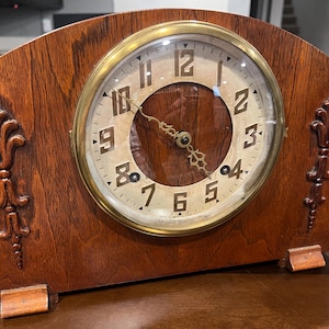 Puede incluir: Un reloj de sobremesa de madera vintage con una esfera de color claro y adornos dorados. El reloj presenta una caja de madera marrón oscuro con tallas decorativas a cada lado. La esfera del reloj muestra números y manecillas negras.