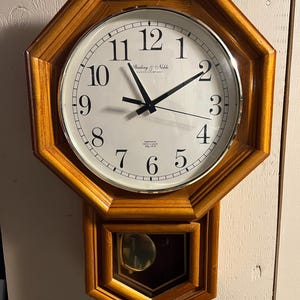 May include: A wooden wall clock with an octagonal frame. The clock face is white with black numerals and hands. The clock has a pendulum in a rectangular frame below the clock face. The text "Sterling & Noble Clock Company" is visible.