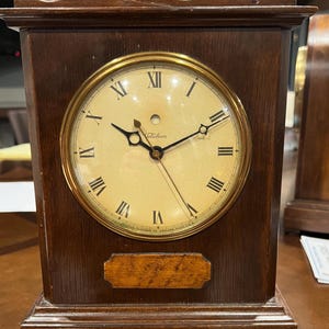 以下が含まれることがあります： 金色のベゼルとローマ数字の目盛りが特徴の、木製のヴィンテージTelechron時計。 時計は長方形で、装飾的なトップフィニアルが付いています。 時計の文字盤はクリーム色です。