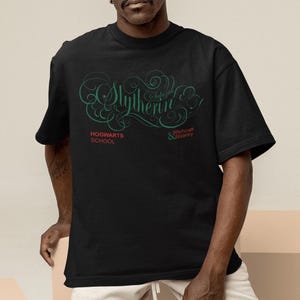 May include: Black t-shirt with the word "Slytherin" in a green, stylized font. The shirt also features the text "Hogwarts School" and "Witchcraft & Wizardry" in green. The shirt is a short-sleeved crew neck.