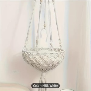 May include: A milk white macrame plant hanger with a woven basket design. The plant holder is suspended by four cords with wooden beads and a tassel at the bottom. The text "Color: Milk White" is at the bottom.