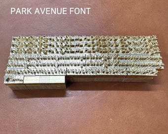 May include: A set of brass letterpress printing blocks arranged in rows. The blocks are arranged alphabetically and numerically. The blocks are on a brown leather surface. The text "PARK AVENUE FONT" is visible at the top of the image.