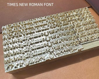 May include: A set of metal letter stamps in a Times New Roman font. The stamps are arranged in rows and columns, with each letter or symbol in its own individual compartment. The stamps are made of a shiny, gold-colored metal.
