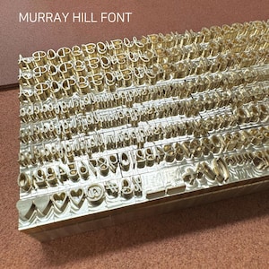 Könnte beinhalten: Nahaufnahme einer Sammlung von Messingstempeln mit Buchstaben, Zahlen und Symbolen, die in Reihen angeordnet sind. Der Text "MURRAY HILL FONT" ist oben sichtbar. Die Stempel werden zum Prägen von Designs verwendet.