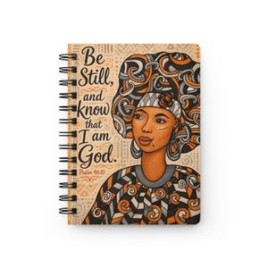 Pode incluir: Um caderno de espiral com a ilustração de uma pessoa com um lenço grande e estampado em tons de laranja, preto e branco. O texto "Be Still, and know that I am God. Psalm 46:10" está impresso no lado esquerdo da capa do caderno.