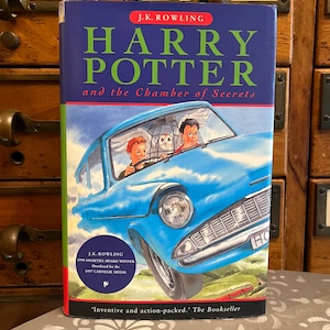 May include: A hardcover book titled "Harry Potter and the Chamber of Secrets" by J.K. Rowling. The cover features a blue car with Harry and Ron, and an owl. The book has a blue background with red and green accents.