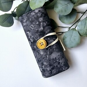 May include: A black fabric pencil case with a floral pattern and a wooden button closure. The case is rolled up and tied with a white ribbon.