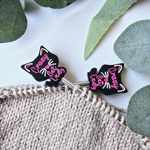 May include: Two black cat-shaped knitting stitch markers with pink lettering that reads "Crazy Cat Lady". The markers are on a beige knitted fabric.